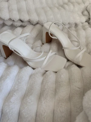 Klackar  - Säljer ett par stilrena vita pumps med fyrkantig tå, blockklack och tunna remmar som korsar foten. Skorna har en modern design och är tillverkade i syntetmaterial. Perfekta för dig som gillar trendiga och minimalistiska skor. Använt en gång🤍