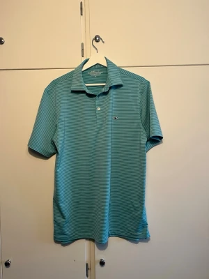 Randig blå polotopp  - Snygg blå polotopp från Vineyard Vines med tunna vita ränder. Klassisk krage, korta ärmar och knappslå med två knappar. Tillverkad i ett mjukt och lätt material som passar perfekt till sommaren. Liten logga på bröstet för en clean look. Perfekt för golf och allmänt varma dagar. 