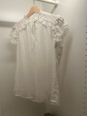 Vit prickig blus med volanger Tamara H. - Söt vit blus från Tamara H. med svarta små prickar, volangdetaljer vid axlar och hals samt knytband fram. Blusen har korta puffärmar och är gjord i ett tunt, lätt material som känns luftigt och somrigt.