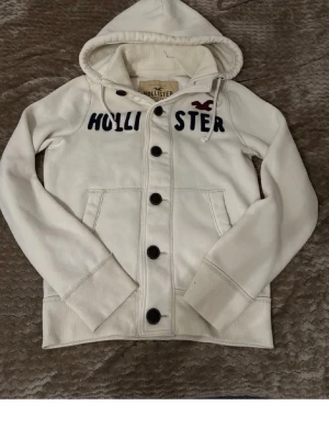  jacka från Hollister  - Snygg vit hoodie från Hollister med stora svarta knappar framtill och broderad logga i marinblått och vinrött på bröstet. Hoodien har huva med dragsko och två fickor framtill. Perfekt för en avslappnad och trendig stil.