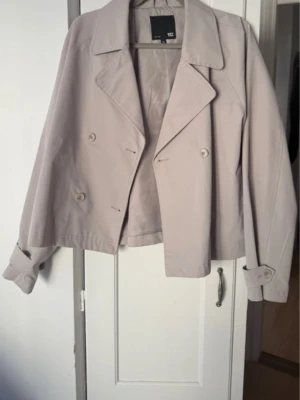 Kort beige trenchcoat från lager 157 - Stilren och kort trenchcoat från lager 157 i ljusbeige färg. Jackan har dubbelknäppning, klassisk krage och dekorativa knappar vid ärmslut. Materialet är slätt och lätt, perfekt för lager-på-lager. Snygg och enkel att matcha med olika outfits.