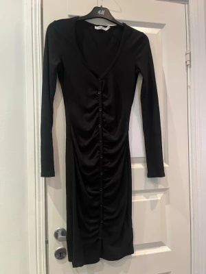 Svart långärmad klänning nakD - Svart långärmad klänning från H&M med v-ringning och knappar framtill. Klänningen har en åtsittande passform och rynkade detaljer längs knappraden. Perfekt för dig som gillar en enkel men stilren look.