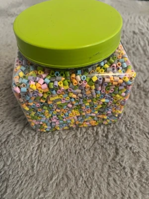 Burk med pastellfärgade pärlor IKEA - Stor plastburk med pastellfärgade rörpärlor från IKEA. Pärlorna är i färger som ljusblå, rosa, gul, grön och lila. Perfekt för pyssel och kreativa projekt. Burken har ett grönt lock och är transparent så man ser pärlorna. Har nog aldrig använt dessa tror jag. 