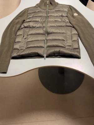 Olivgrön dunväst från Moncler - Snygg olivgrön dunväst från Moncler med stickade ärmar och hög krage. Västen har quiltad front, två fickor med dragkedja och en Moncler-logga på ärmen. Perfekt för lager-på-lager och ger en sportig vibe till din outfit.