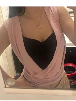 Superfin topp💕 - Säljer en ljusrosa omlott topp med djup v-ringad urringning och draperad front. Toppen är ärmlös och har en snygg croppad passform som framhäver midjan. Under finns en svart bandeau som är fastsytt på den rosa ger en cool kontrast. Perfekt för dig som gillar trendiga och unika plagg. Riktig vintage y2k