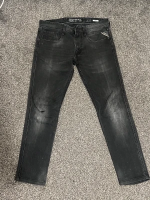 Svarta slim fit-jeans från Replay  - Säljer ett par svarta slim fit Replay anbass  med snygga slitningar och klassisk femficksdesign. Jeansen har diskreta detaljer och en liten logotyp på myntfickan. Perfekta för en trendig och avslappnad stil.