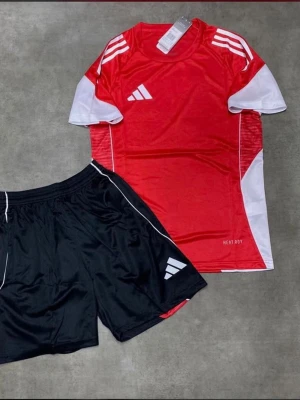 Adidas röd och vit träningsset - Snyggt träningsset från Adidas med en röd och vit t-shirt och svarta shorts. T-shirten har vita detaljer på axlar och sidor samt Adidas-logga på bröstet. Shortsen är svarta med vit logga och tunn vit linje längs sidan. Perfekt för fotboll eller gym.