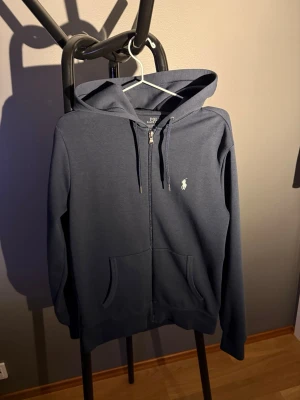 Mörkblå hoodie från Polo Ralph Lauren - Mörkblå hoodie med dragkedja från Polo Ralph Lauren. Klassisk design med huva, dragsnören och två fickor framtill. Ikoniska broderade loggan på bröstet. Perfekt för en avslappnad och stilren look.