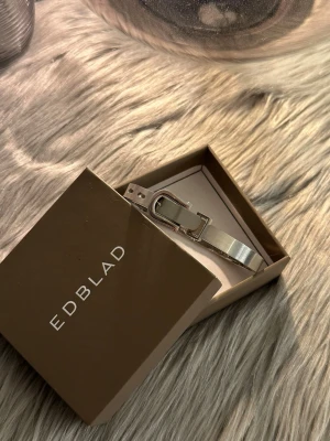 Silverfärgat armband från Edblad - Snyggt armband i silverfärgat rostfritt stål från Edblad. Armbandet har en unik design med ett spänne som liknar ett bälte, vilket ger en cool och modern look. Använd ett par gånger  och är i nyskick. Orginallåda medföljer