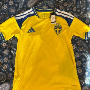 Sverige matchtröja Adidas gul/blå - Gul matchtröja från Adidas med Sveriges landslagsemblem på bröstet och blå detaljer på axlar och ärmar. Tröjan har ClimaCool-material för bra ventilation och komfort. Perfekt för fotbollsträning eller att bära på matchdagar. Storlek S