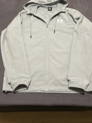 Ljusgrå vindjacka från Under Armour 💯% äkta från zalando  - Snygg ljusgrå vindjacka från Under Armour med dragkedja framtill, huva med dragsko och elastiska muddar vid ärmsluten. Jackan har en minimalistisk design med logga på bröstet och är tillverkad i ett lätt, vindtätt material.