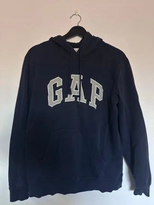 Mörkblå hoodie från GAP - Mörkblå hoodie från GAP med stor logga i vitt och grått på bröstet. Klassisk modell med huva och dragsko samt en stor magficka framtill. Tillverkad i mjukt bomullsmaterial som är skönt att ha på sig. Perfekt för en avslappnad och stilren look.