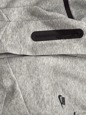 Grå Nike Tech Fleece mjukisbyxor - Nike Tech Fleece mjukisbyxor i ljusgrå färg med svarta detaljer och logga. Byxorna har en modern passform, dragkedjeförsedd sidoficka och är tillverkade i mjukt fleecematerial. Perfekta för chill eller träning.KAN ÄVEN KÖPAS SEPARAT