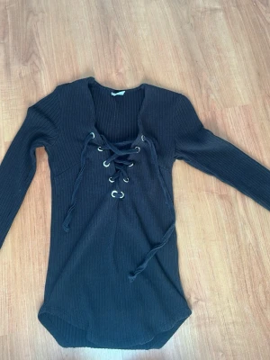 Svart ribbad stickad tröja med snörning - Säljer en svart, ribbad stickad tröja med långa ärmar och snygg snörning framtill. Tröjan har rund halsringning och metallöljetter vid snörningen för en cool detalj. Perfekt för dig som gillar trendiga och bekväma plagg.