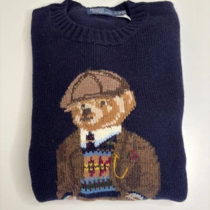 Mörkblå stickad tröja med björn - Säljer en mörkblå stickad tröja från Polo Ralph Lauren med en stor broderad björn på framsidan, klädd i klassisk outfit med kavaj, väst och keps. Tröjan har rund halsringning och ribbade muddar. Perfekt för dig som gillar preppy stil och detaljer.