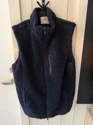 Mörkblå teddyväst - Mysig mörkblå teddyväst från Cubus Design med hög krage och hel dragkedja framtill. Västen har en bröstficka med dragkedja och är tillverkad i fluffigt teddy-material som håller dig varm. Perfekt för lager-på-lager-stil.