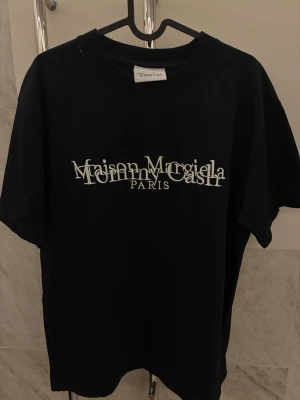 Maison Margiela x Tommy Cash svart t-shirt - Svart t-shirt med Maison Margiela x Tommy Cash Paris-tryck i vitt framtill. Klassisk rund hals och korta ärmar. Oversized passform och tillverkad i mjuk bomull. Perfekt statement-plagg för dig som gillar unika samarbeten.