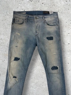 Ljusblå jeans med slitningar - Säljer ett par ljusblå jeans med flera slitningar och hål på benen. Jeansen har fem fickor, smal passform och klassisk knappgylf. Perfekta för en avslappnad och trendig look. W30 Slim Glenn