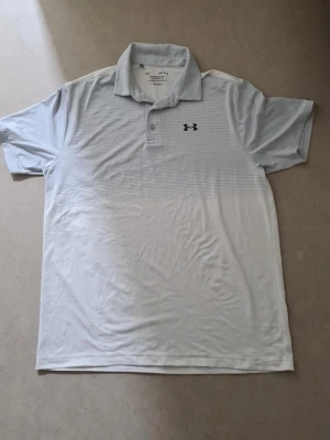 Vit piké från Under Armour - Säljer en stilren vit pikétröja från Under Armour med diskreta horisontella ränder upptill och klassisk krage. Tröjan har korta ärmar och knappar vid halsen. Perfekt för en sportig och avslappnad look.