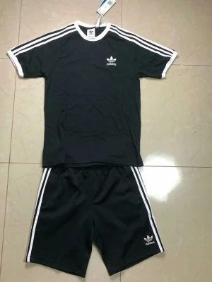 Svart Adidas träningsset med vita ränder - Snyggt svart träningsset från Adidas med klassiska vita ränder längs ärmar och ben. Setet består av en t-shirt och shorts, båda med Adidas-logga på bröstet och benet. Perfekt för träning eller sportiga dagar. Materialet är mjukt och bekvämt.