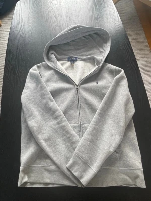 Grå hoodie från Polo Ralph Lauren - Snygg grå hoodie med dragkedja från Polo Ralph Lauren. Klassisk design med broderad logga på bröstet, huva och mjukt fleecefoder på insidan. Perfekt för en chill och avslappnad stil.                Storlek: XS, det står XL men det är i barn så den passar XS i vuxen📏                                             Frakt: Billig och snabb frakt 🚚 💴 