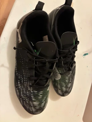Svarta Puma fotbollsskor med mönster - Säljer ett par svarta fotbollsskor från Puma med vitt och grönt mönster på ovandelen. Skorna har klassisk snörning, låg profil och vita dobbar för bra grepp på planen. Tillverkade i syntetmaterial för lätt känsla och smidig passform.
