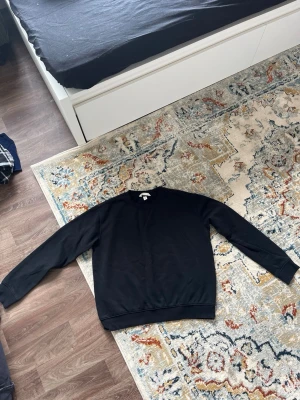 Svart sweatshirt från H&M - En stilren svart sweatshirt från H&M med rund halsringning och ribbade muddar vid ärmslut och nederkant. Perfekt för en avslappnad look och enkel att matcha med andra plagg. Tillverkad i mjukt bomullsmaterial.