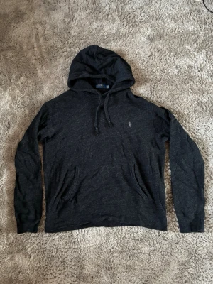 Mörkgrå hoodie från Polo Ralph Lauren - Mörkgrå hoodie från Polo Ralph Lauren med klassisk huva och dragsnören. Diskret logga broderad på bröstet och praktisk magficka. Materialet är mjukt och bekvämt, perfekt för en avslappnad stil. Långärmad och stilren design.