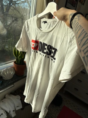 Vit Diesel t-shirt med tryck - Säljer en vit t-shirt från Diesel med stort tryck i svart och rött på bröstet där det står 'Diesel Denim Division'. Klassisk passform och rund hals. Perfekt för dig som gillar streetwear och vill ha en enkel men snygg look.