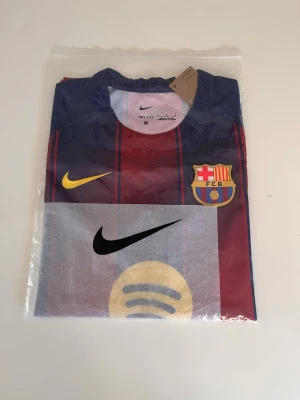 FC Barcelona matchtröja Nike - Säljer en officiell FC Barcelona matchtröja från Nike med klubbmärke, gula och svarta Nike-loggor samt Spotify-logga på bröstet. Tröjan har marinblå och vinröda ränder och är tillverkad i lätt polyester, perfekt för fotbollsträning eller match.
