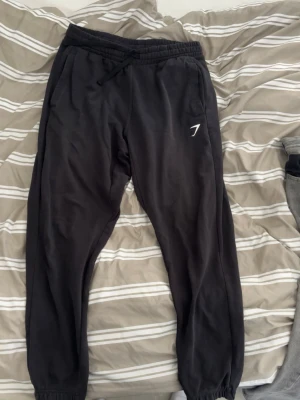 Svarta mjukisbyxor från Gymshark - Säljer ett par svarta mjukisbyxor från Gymshark med dragsko i midjan och resår vid bensluten. Byxorna har en enkel och clean look med Gymshark-logga på vänster ben. Perfekta för chill eller träning.