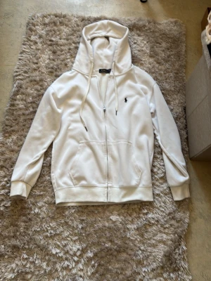 Vit hoodie från Polo Ralph Lauren - Säljer en vit hoodie från Polo Ralph Lauren med dragkedja framtill, två fickor och den klassiska lilla loggan broderad på bröstet. Hoodien har justerbar huva med snören och ribbade muddar vid ärmar och nederkant. Perfekt för en clean och stilren look.