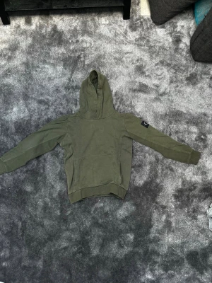 Olivgrön hoodie från Calvin Klein - Snygg olivgrön hoodie från Calvin Klein med klassisk huva och ribbade muddar. På vänster ärm sitter en svart patch med CK-logga. Perfekt för en avslappnad och trendig stil. Materialet känns mjukt och bekvämt. Storlek 14