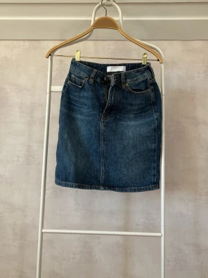 Jeanskjol från Samsøe Samsøe - Snygg blå jeanskjol från Samsøe Samsøe i klassisk modell med fem fickor och knappgylf. Kjolen har en rak passform och är tillverkad i slitstarkt denimtyg. Perfekt att matcha med en enkel t-shirt eller stickad tröja. (Aldrig använd, men saknar prislapp)