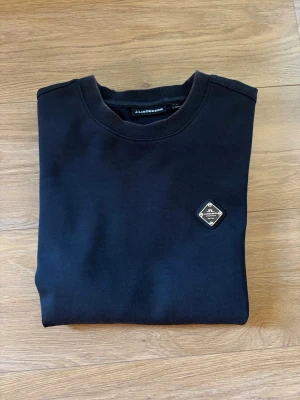 J.Lindeberg tröja - En helt vanlig svart tröja från J.Lindeberg med en metall logga på vänster bröst. Storlek S, bra skick, inga defekter. Lite solblekt på kanterna av tröja. Köpte den för 1400kr för något år sedan, tror inte man kan köpa den längre. Skriv vid frågor!