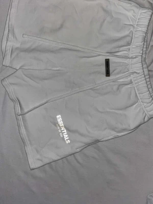 Vita Essentials shorts med tryck - Säljer ett par vita shorts från Essentials med tryck på benet och elastisk midja. De har en avslappnad passform och är tillverkade i mjuk bomull, perfekta för sommardagar. Klassisk och enkel design med diskret logga.