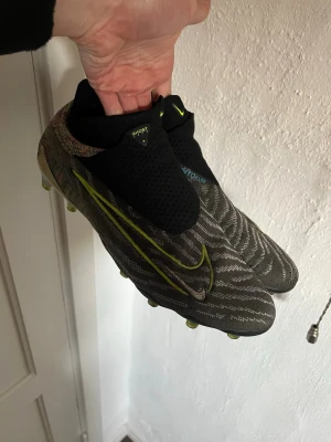 Nike Phantom Vision Ghost Lace FG - Säljer ett par Nike Phantom Vision Ghost Lace fotbollsskor med FG-dobbar. Skorna är svarta och grå med mönstrad ovandel, gula detaljer och en stor Nike Swoosh på sidan. Ovandelen är i syntetmaterial och har ett strumpliknande skaft för extra komfort.