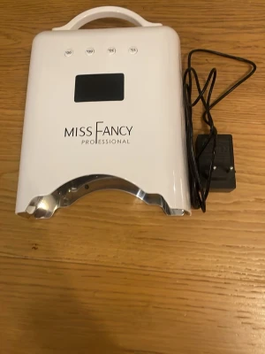 Miss Fancy UV-lampa för naglar - UV-lampa från Miss Fancy Professional i vit plast med digital display och fyra timerknappar. Perfekt för att härda gelélack och andra nagelprodukter. Lampan har en smidig bärhandtag och medföljer strömadapter. Modern och stilren design. Nypris 1299kr.