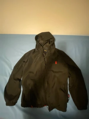 Svart vindjacka från Polo Ralph Lauren - Svart vindjacka med huva från Polo Ralph Lauren. Jackan har dragkedja framtill, två sidofickor och den klassiska röda Polo-loggan på bröstet. Materialet är lätt och vattenavvisande, perfekt för blåsiga dagar.
