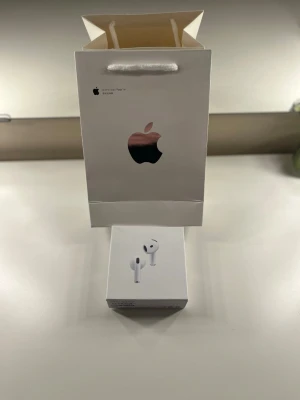 Apple AirPods 4 - Säljer ett par vita Apple AirPods med trådlöst laddningsetui. Hörlurarna har en stilren och minimalistisk design i blank plast och levereras i originalförpackning med tillhörande manual. Perfekta för musik, samtal och vardagsbruk.