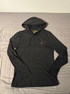 Svart hoodie från Polo Ralph Lauren - Svart hoodie från Polo Ralph Lauren med klassisk huva och dragsko. Långärmad modell i mjukt och tunt material, perfekt för en avslappnad stil. Ikoniska röda Polo-loggan broderad på bröstet ger en snygg detalj.