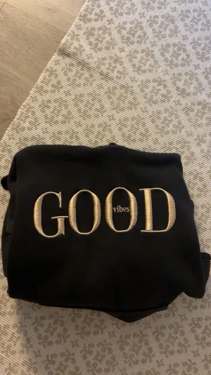 Svart hoodie med brodyr GOOD vibes - Svart hoodie med beige broderad text 'GOOD vibes' på bröstet. Hoodien har huva med beige snören och en stor magficka framtill. Perfekt för en chill och avslappnad stil.