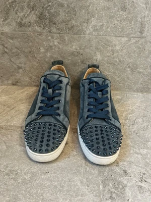 Blå sneakers med nitar från Louboutin - Snygga blå sneakers från Christian Louboutin med coola blå nitar på tån och klassisk röd sula. Skorna har blå snörning och är gjorda i mocka och läder. Perfekta för dig som vill sticka ut med en edgy stil. Kan gå ner i pris vid snabb affär.
