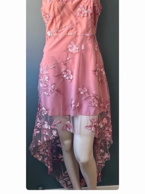 Rosa blommig långklänning med spets - Romantisk rosa långklänning med transparent spets och broderade blommor. Klänningen har asymmetrisk nederkant och vackra detaljer som ger en lyxig känsla. Perfekt för speciella tillfällen när du vill sticka ut.