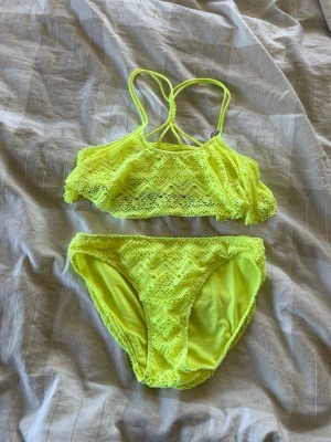 Gul volangbikini med spetsdetaljer - Snygg gul bikini med volang och spetsdetaljer. Överdelen har smala axelband som korsas i ryggen, en söt volangkant samt en silver löv detalj vid ena axelbandet. Underdelen har matchande spetsmönster och normal passform.