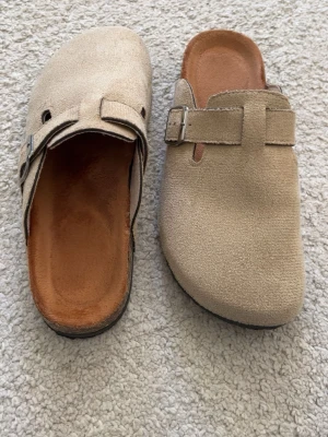 Birkenstock tofflor - Säljer ett par snygga beige tofflor i mocka med justerbart spänne i metall. De har en klassisk slip-in design och en bekväm innersula i brunt. Perfekta för dig som gillar stilrena och tidlösa skor.