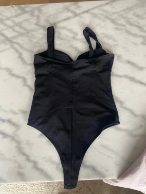 Svart body med breda axelband - Snygg svart body med breda axelband och lätt vadderade kupor. Modellen har en markerad midja och hög skärning vid benen. Materialet ser ut att vara stretchigt och följsamt, perfekt för att skapa en smickrande siluett.