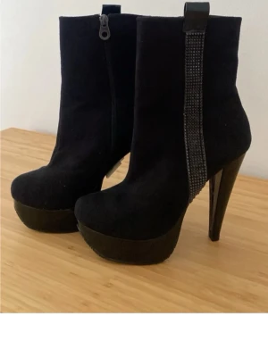 Svarta platåstövletter med strass - Snygga svarta stövletter med hög platå och smal klack. Dekorativ strassdetalj längs sidan och dragkedja på insidan. Tillverkade i mockaimitation för en lyxig känsla. Perfekta för dig som vill sticka ut med höga klackar och cool design.
