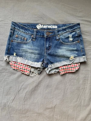 Blå jeansshorts med rutiga detaljer - Snygga blå jeansshorts från Hermosa med slitna detaljer och råa kanter. Under shortsen syns röda och vita rutiga tygdetaljer som sticker ut. Klassisk femficksmodell med knapp och dragkedja framtill. Perfekta för en avslappnad stil.