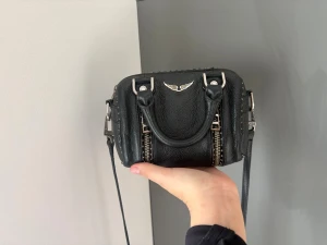Svart handväska från Zadig & Voltaire - Säljer en svart handväska från Zadig & Voltaire i strukturerat läder med silverfärgade metalldetaljer och två dragkedjor framtill. Väskan har både handtag och en lång axelrem, samt den ikoniska Zadig & Voltaire-loggan i metall på framsidan. Skicket är ändå hyfsat bra men lite rostig på några ställen som inte jag skulle säga märks av mycket. Köpt i matbella för 275 euro säljer för 1500